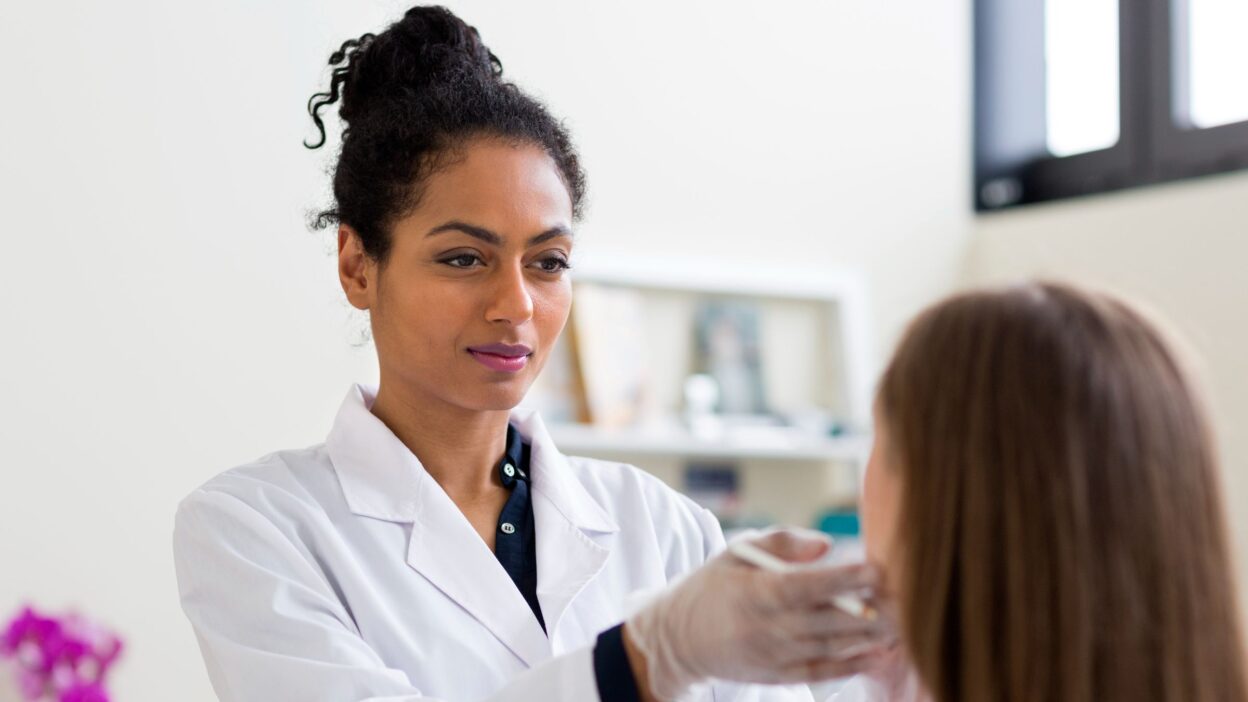 A clinician wearing a white coat and gloves examines a patient’s face during a consultation.