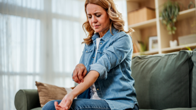 Woman sitting on a couch scratching her itchy forearm, wincing in discomfort